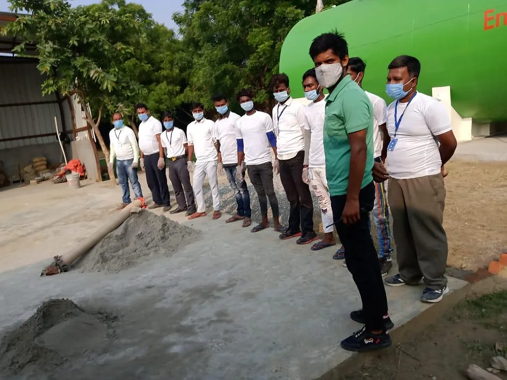 A line of field workers in white t-shirts, masks, gloves and ID cards at a processing site with a large green tank behind them