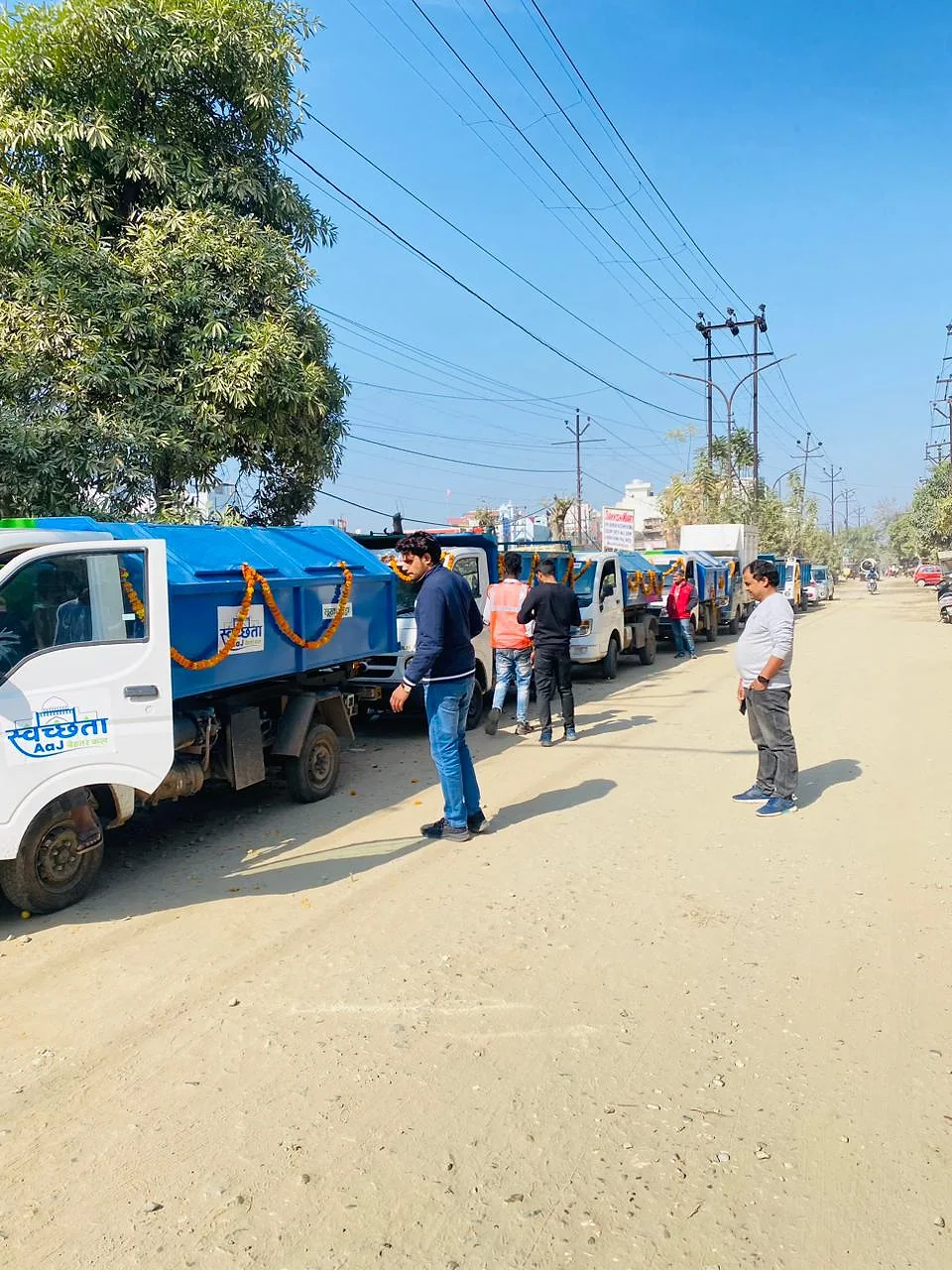 A row of blue waste collection tippers decorated with marigold garlands, parked along a sunny road with team members beside them