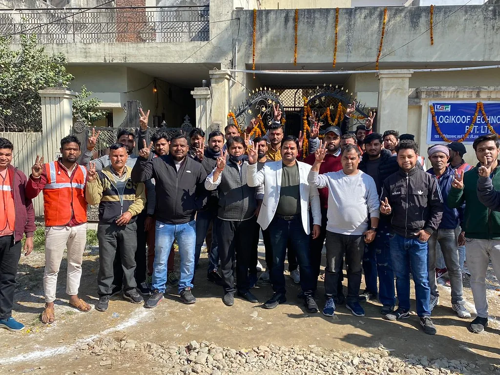 A large group of team members, some in high-visibility vests, raising victory signs outside a building decorated with marigolds